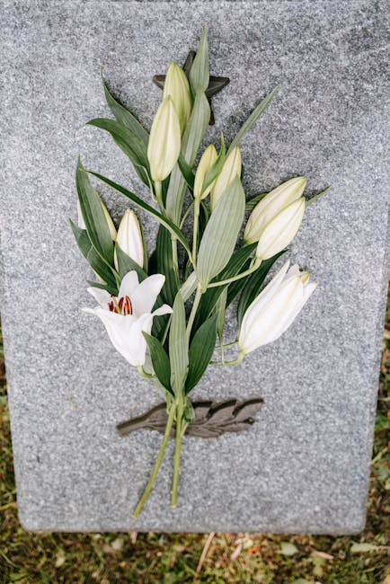 A delicate floral arrangement featuring white lilies and white tulips with closed buds, arranged with green foliage and long, slender leaves. The flowers are positioned against a grey stone surface, s