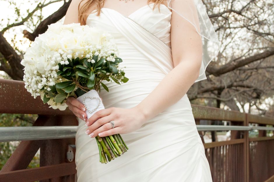 A bride holding a bouquet of fresh white flowers, including roses and baby's breath, arranged with dark green foliage and wrapped in white paper with a decorative heart-shaped clasp. The bouquet is po