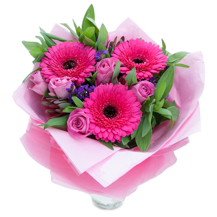 I see a bouquet that balances boldness with tenderness: three radiant fuchsia Gerbera daisies dominate the scene, each composed of many narrow petals that fan out from a deep, dark centre, creating energetic focal points. Surrounding these are several dusty lavender-pink roses, their petals curved inward in the early stages of bloom, offering soft, velvety layers that invite a gentle touch. Tiny purple statice flowers are tucked among the larger blooms, their papery texture and cool hue adding depth and a meadow-like sparkle that offsets the saturated pinks. The greenery is abundant and thoughtfully placed - broad, glossy leaves that resemble salal and whispery eucalyptus branches build a living backdrop, giving the bouquet fullness and a faint, green aroma. The presentation is as deliberate as the floristry: an inner wrap of pale pink, matte tissue provides a cushioned foundation, while a shimmering, semi-translucent fuchsia-pink cellophane envelops the arrangement with slight crinkles that catch the light and unify the colour story. The wrapping is artfully folded, suggesting care taken to prepare the bouquet for gifting or bedside display; a clear glass vase's rim is visible at the lower edge, implying immediate readiness for water. This composition feels perfectly at home beside a bookshop window on Church Road in Barnes or brightening a living room after a stroll around Barnes Common. It carries a light perfume of roses and an understated freshness that makes it ideal for celebrating birthdays, Mother's Day, anniversaries, or a quiet, uplifting surprise.